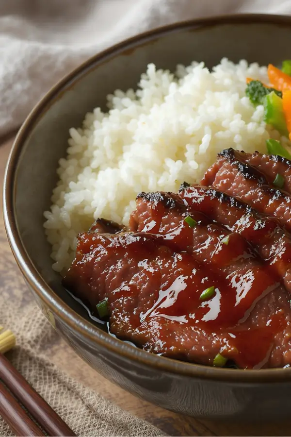Thin sliced caramelized beef glazed with shiny teriyaki sauce, served with steamed white rice and stir-fried vegetables on a ceramic bowl